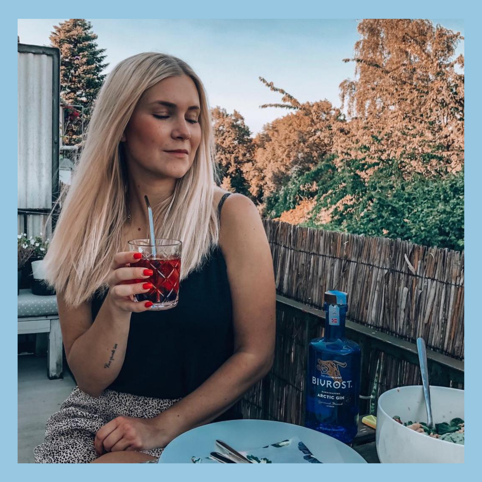Eine blonde Frau sitzt mit einem Getränk auf einem Balkon.